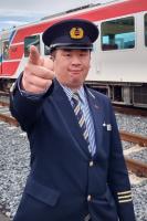 停車する列車の前で、鉄道会社の制服を着て人差し指で前方を差す男性を正面から写した写真