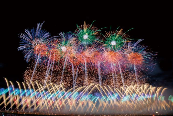 夜空に色とりどりの大輪の打ち上げ花火が打ち上がり、その下から複数の花火が交差になり夜空に上がっている花火の写真