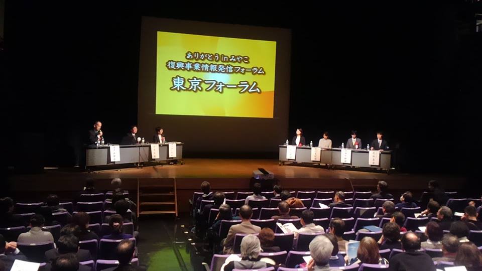 画面が映されたスクリーンの左右に並べられたテーブルの席につく関係者の方々と、会場の椅子に座り舞台上を見ている参加者の方々の写真