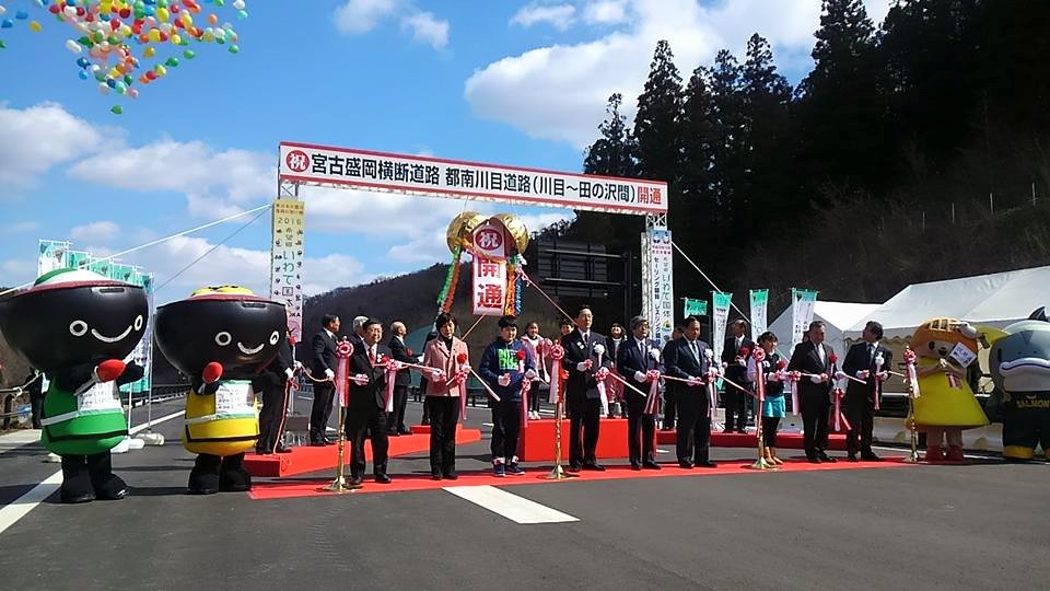 道路を横断するように敷かれたレッドカーペットの上でテープカットをする関係者の方々と、わんこ兄弟、サーモンくん、開運かなえちゃんの写真