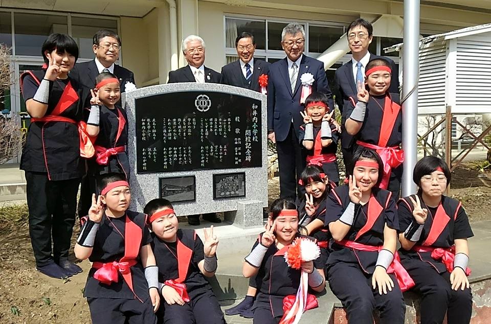 石碑の周りに集まる太鼓の衣装を着た小学生たちと、関係者の男性たちの記念写真