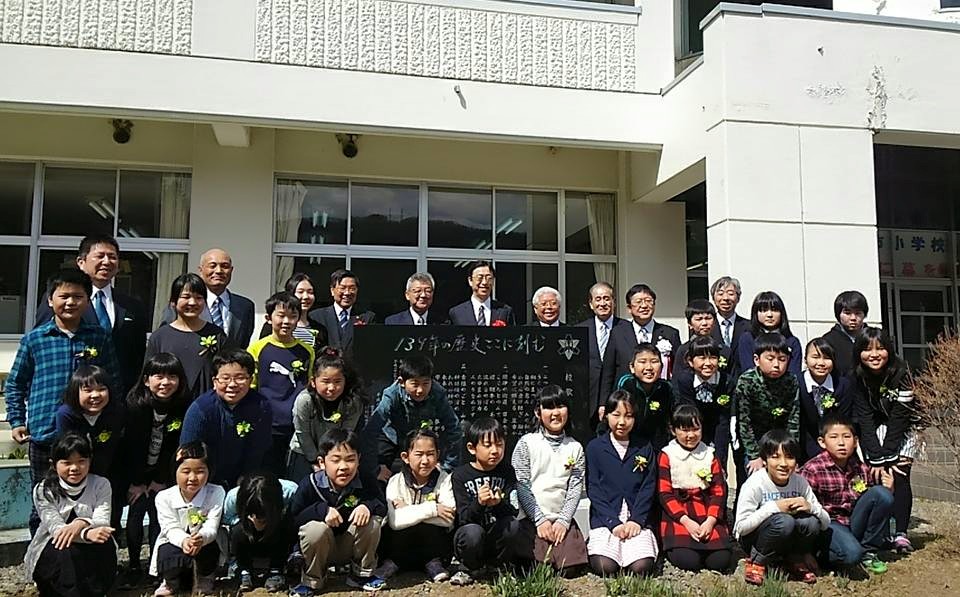 石碑の周りにしゃがんだり中腰になったり立って並んでいる小学生たちと、関係者の方々の記念写真