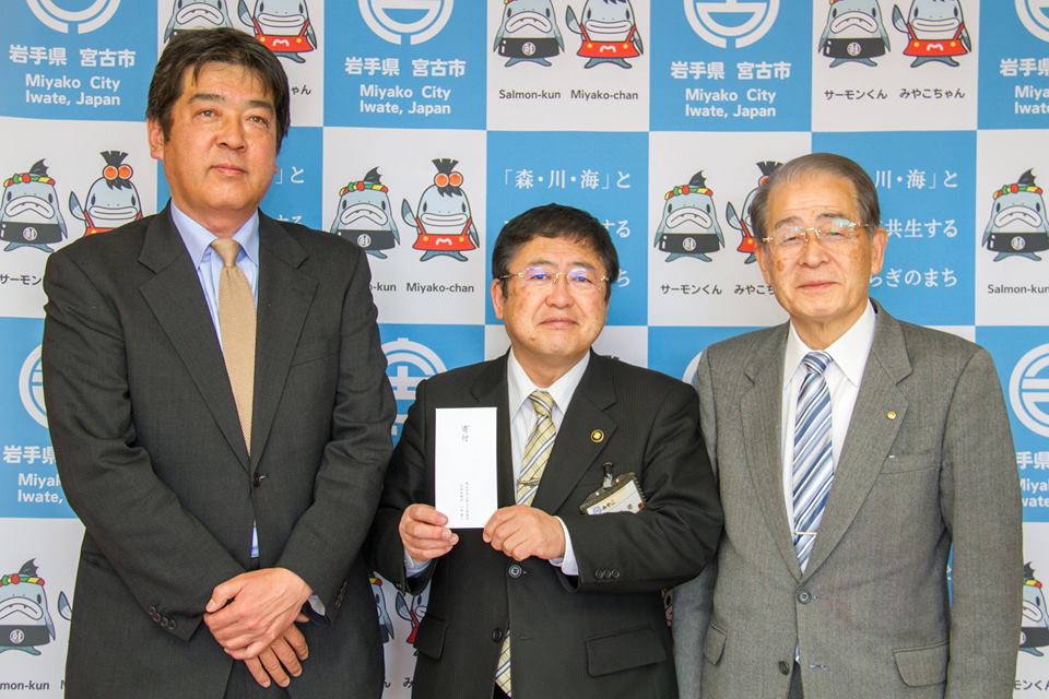 バックパネルの前で寄附金の封筒を持つ市長を中心に、男性2名が両側に立っている記念写真