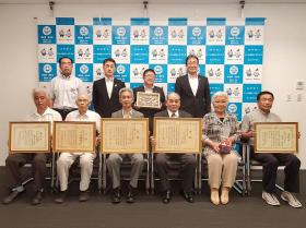 バックパネルの前に「全農乾椎茸品評会」を受賞した方が賞状を手に持ち、横一列に椅子に座って並んでおり、その後ろに市長含めた関係者4名が立ち並んでいる写真
