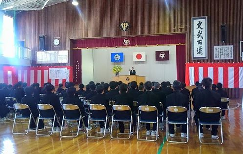 壁に紅白の幕が張られた体育館で、前のステージ上の演台で祝辞を述べている市長と 座って聞いている新入生の写真