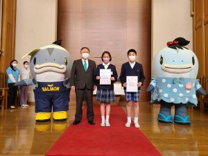 市長と委嘱状を手に持つ子供達と、サーモンくんとみやこちゃんが横に並んでいる写真