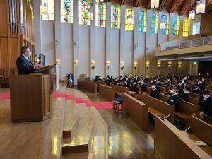 広い礼拝堂の座席に青山学院初等部の6年生の子供達が席に座り、演壇で話をする市長の写真