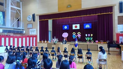 壁には紅白の幕が張られて、入学を祝う花や絵が飾られたステージ下の演台で、 祝辞を述べている市長と、横一列に座って聞いている新一年生と、学年ごとに座っている在校生たちの写真