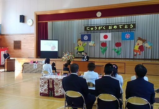 にゅうがくおめでとう書かれた横断幕とお祝いの絵が飾られているステージ下の演台で、祝辞を述べている市長と座って聞いている新一年生、そのご父兄、教員の方々の写真