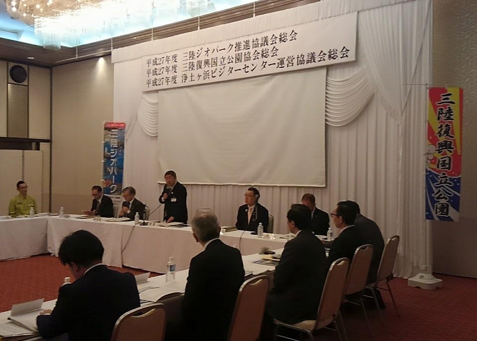 平成27年度三陸ジオパーク推進協議会・三陸復興国立公園協会・浄土ヶ浜ビジターセンター運営協議会総会と書かれたボード下の自席で立って、参加者の方々に説明をしている写真