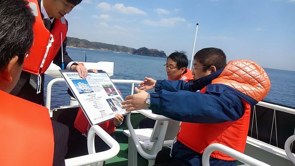 晴天の下、赤のライフジャケットを着た市長が、同乗者と船上で資料を見ながら宮古港を視察している写真