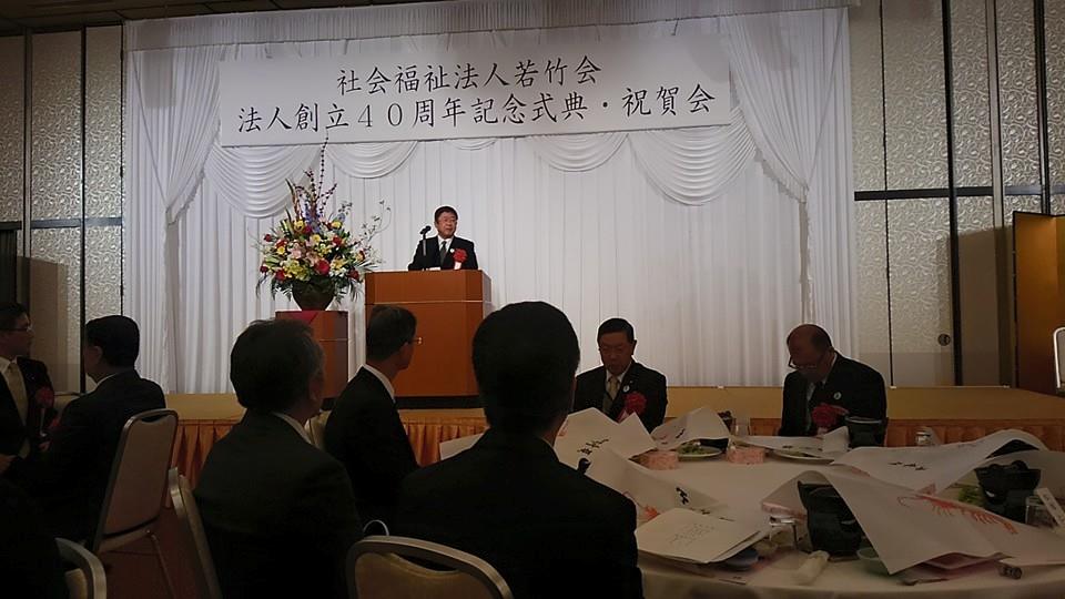 社会福祉法人若竹会法人創立40周年記念式典・祝賀会と書かれた横断幕のステージ上で、 円卓のテーブルに参加者の方々が座っている中、市長が赤の胸章をして、祝辞を述べている写真