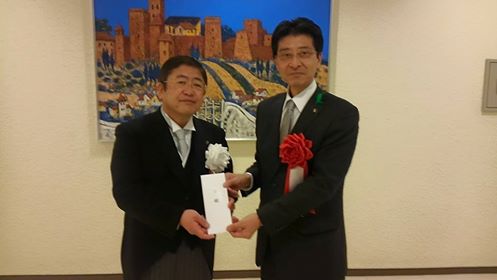 赤の胸章をつけた黒岩市の高樋憲市長から白の胸章をつけた市長へ義援金の目録を渡されている写真