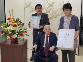 椅子に座った高齢の男性を中心に、熨斗のついた記念品を持つ女性、バインダーを持つ市長が並んでいる記念写真