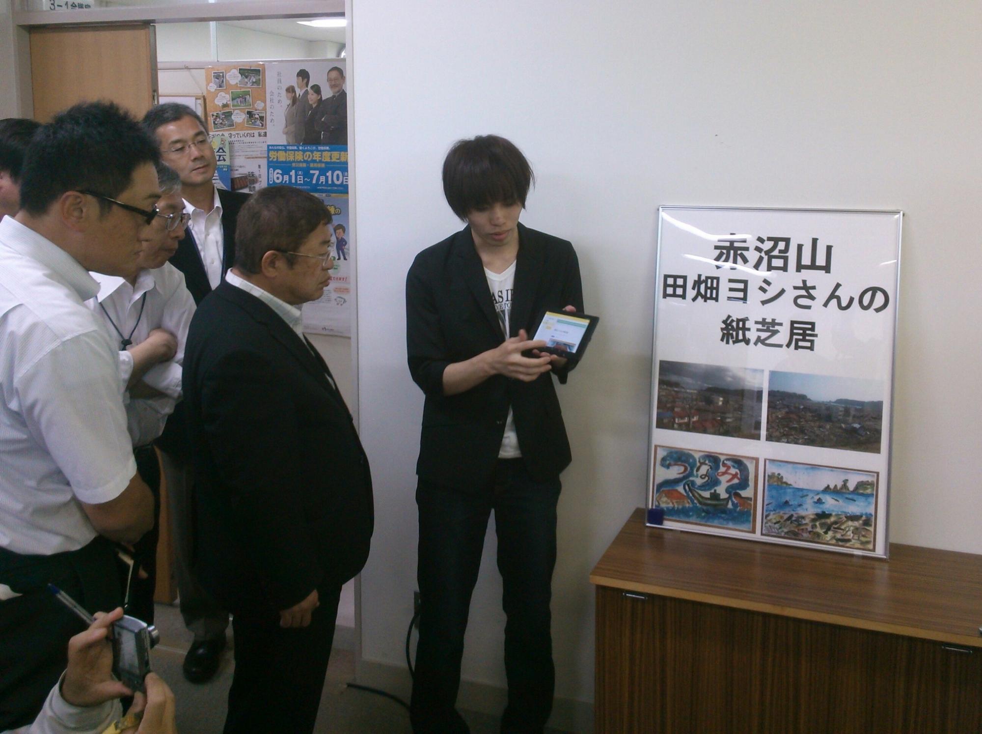 額に入った「赤沼山 田畑ヨシさんの紙芝居」という文字と写真が載ったポスターの横で、小さいモニターを市長に見せている様子の男性の写真
