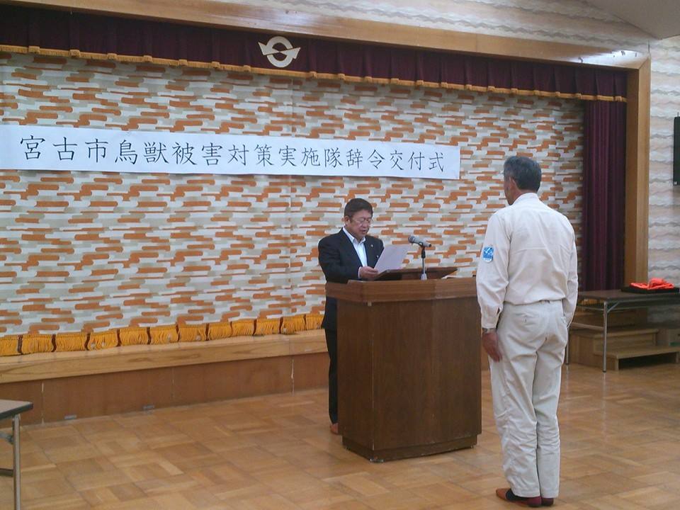 「宮古市鳥獣被害対策実施隊辞令交付式」と書かれたパネルが吊るされた舞台の前で、演台の向かい側に立つ男性にむかって辞令を読み上げている様子の市長の写真
