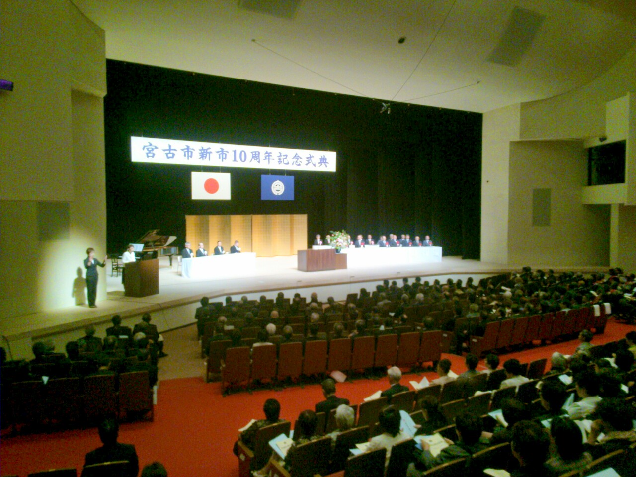 会場の席に座る参加者たちと、舞台上に並べられたテーブルの席に着く関係者の方々の写真