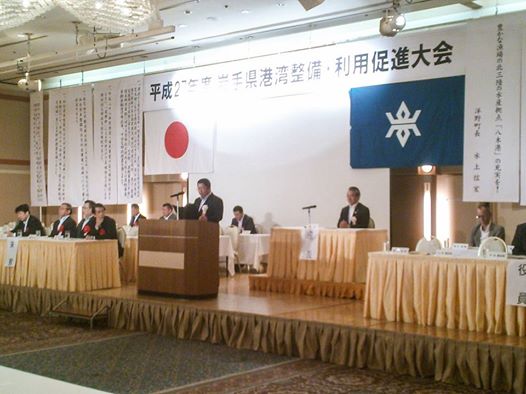 舞台の上に間隔を開けて置かれた机の席に座る関係者の方々と、中央の演台に置かれたマイクに向かい話をする市長の写真