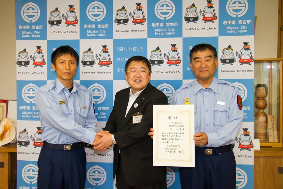 青い消防の制服を着た男性1名と市長と両手で握手をし、隣で賞状を両手で持つ消防の制服を着た男性1名が並ぶ記念写真