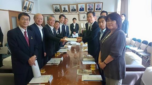 並べられた机に向かい合わせで立つ市長と関係者の方々の中央で要望書を受け渡ししている様子の2名の男性が写った記念写真