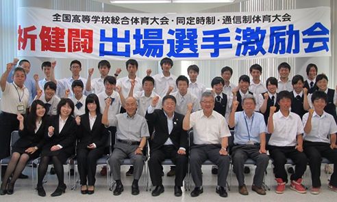 「祈健闘 出場選手激励会」と書かれた横断幕の下で、1列目は椅子に座り、2列に3列目に立って並ぶ市長や出場選手、関係者の方々の記念写真