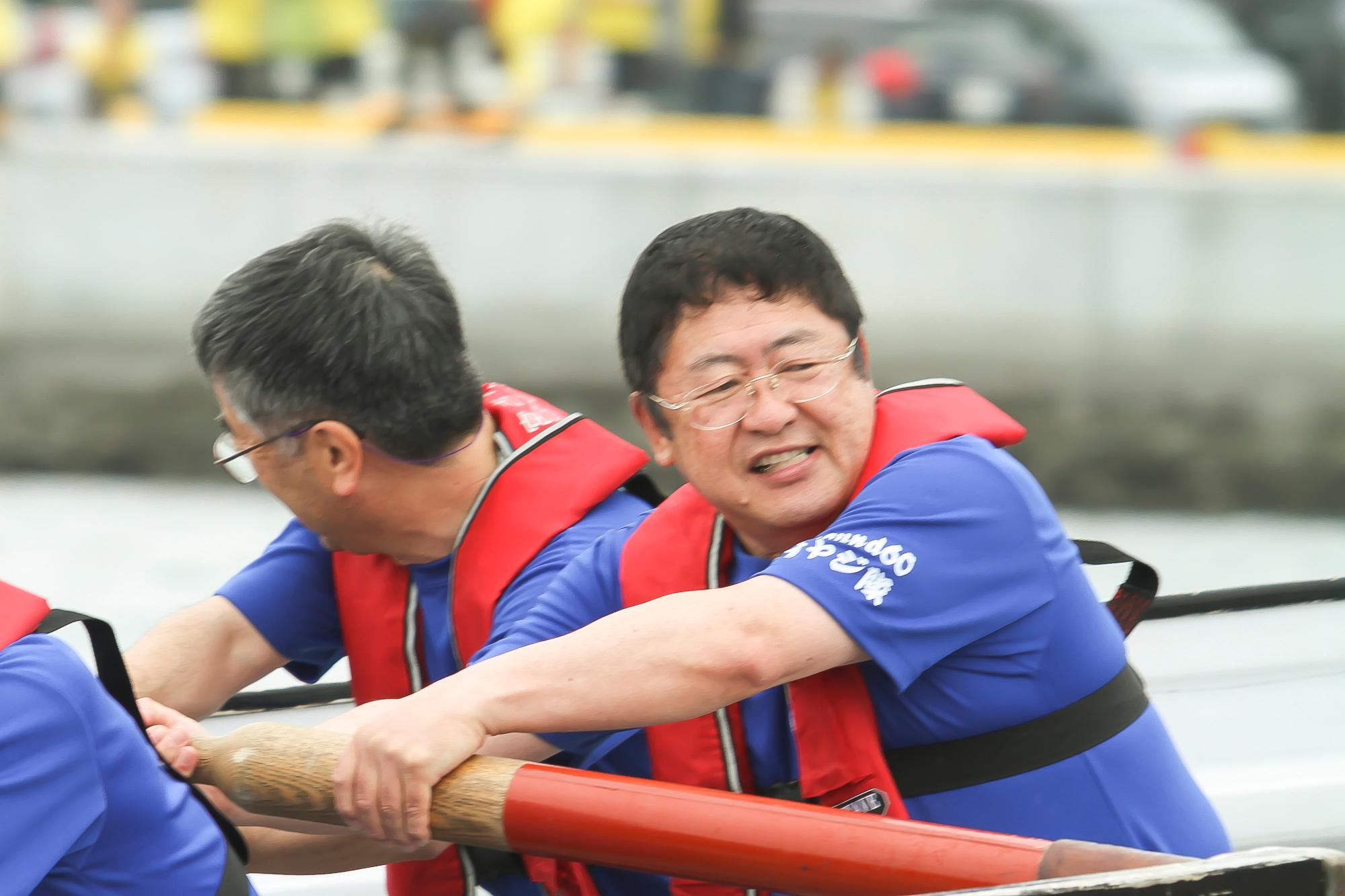 紫色のTシャツを着て赤い救命胴衣を着た市長と男性がカッターに乗り櫂を漕いでいる様子の写真