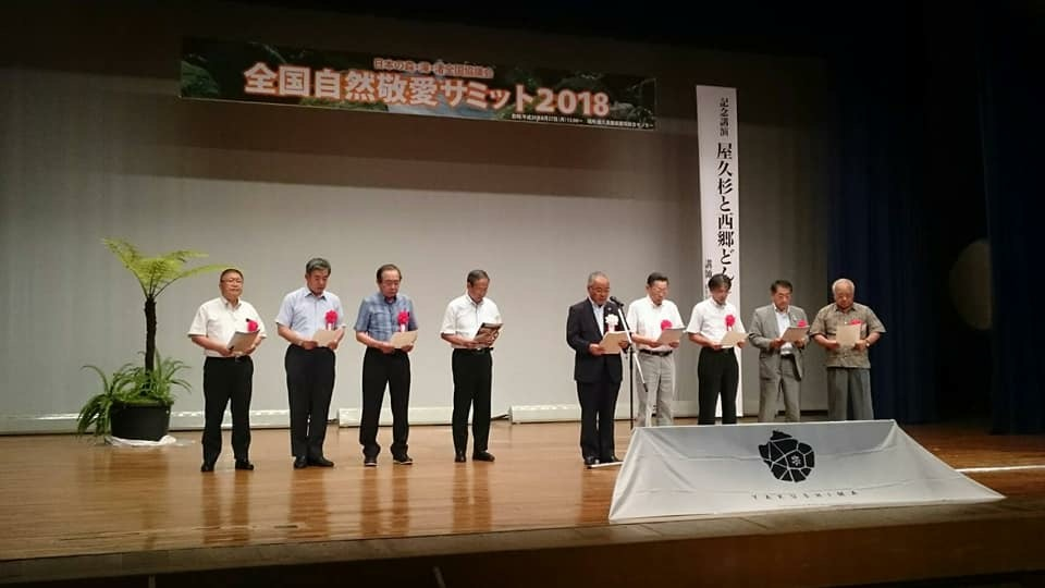 「全国自然敬愛サミット2018」と書かれた横断幕が吊るされた舞台上で、胸章を着けて横一列に並んだ8名の関係者と、中央でスタンドマイクに向かい話をする男性の写真