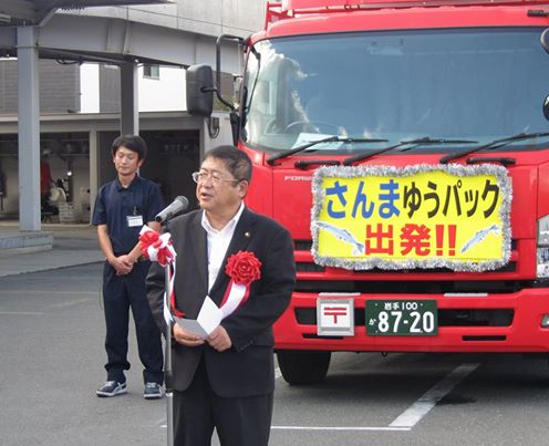 正面に「さんまゆうパック出発!!」と書かれた黄色いパネルが飾られたトラックの前で、紅白リボンがつけられたスタンドマイクに向かい話をする市長の写真
