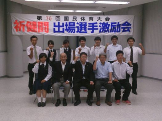 「第70回国民体育大会 祈健闘 出場選手激励会」と書かれた横断幕の下で、椅子に座った学生2名、市長、関係者の男性2名と後列で立つ5名の学生と2名の男性が片方の握りこぶしを顔の横に掲げてポーズをとっている記念写真