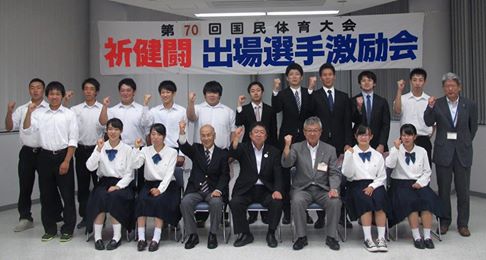 「第70回国民体育大会 祈健闘 出場選手激励会」と書かれた横断幕が掲げられた会場で、選手や市長、関係者の方々が2列に並び右手を上げたポーズをとっている記念写真