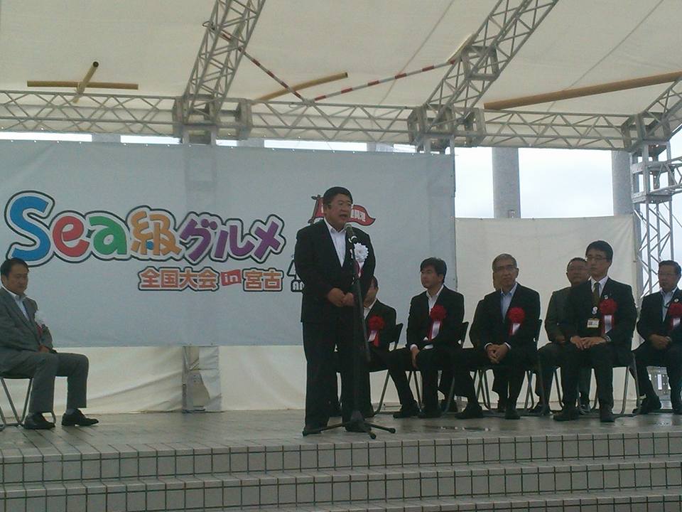 「Sea級グルメ」と書かれた横断幕が掲げられた舞台上で、白い胸章を着けた市長がスタンドマイクに向かい話をする様子の写真