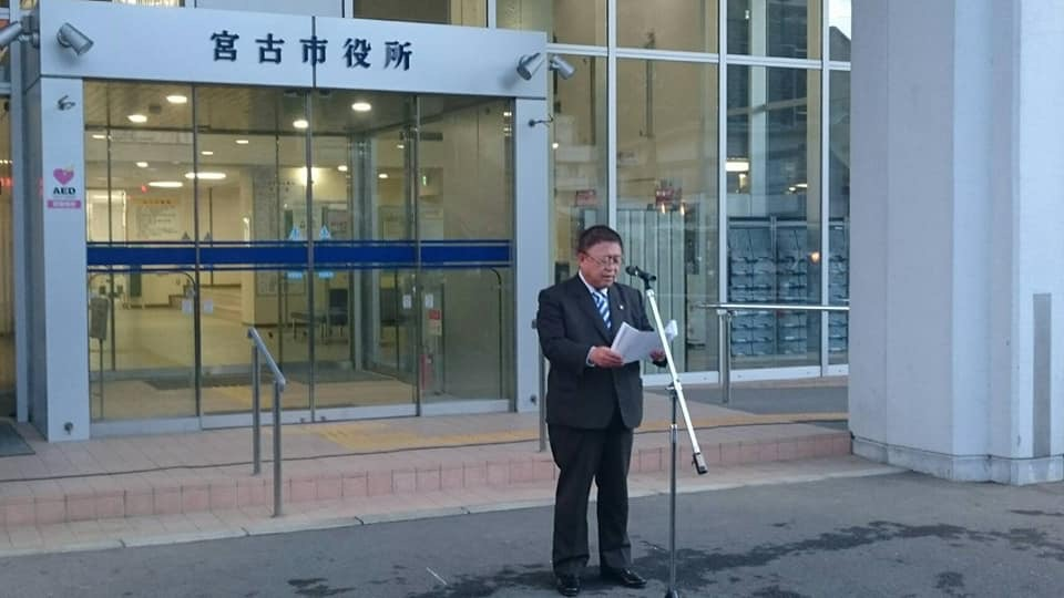 宮古市役所の入り口の前でスタンドマイクに向かい話をする様子の市長の写真