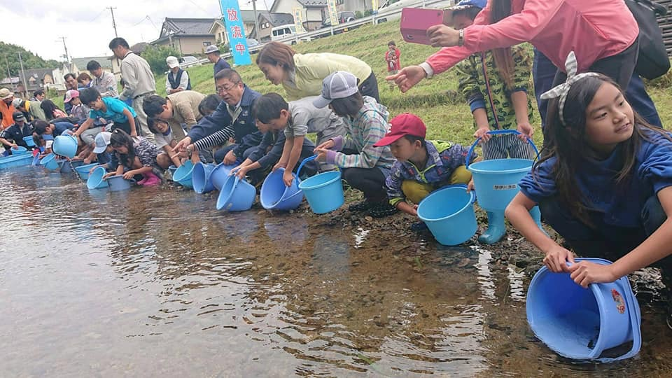 青いバケツを川につけ稚魚を放流している様子の大勢の子供たちと市長の写真