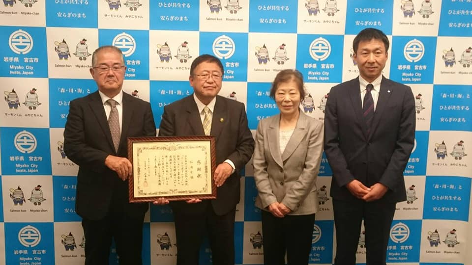 バックパネルの前で、賞状の額縁を一緒に持つ市長と眼鏡をかけた男性、その横に並ぶ女性1名と男性1名の記念写真