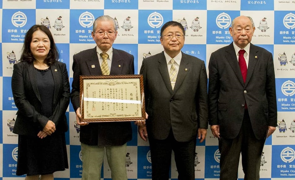バックパネルの前で賞状の額縁を持つ眼鏡をかけた白髪の男性と市長を中心に、女性1名と赤いネクタイをした男性が横一列に並んでいる写真