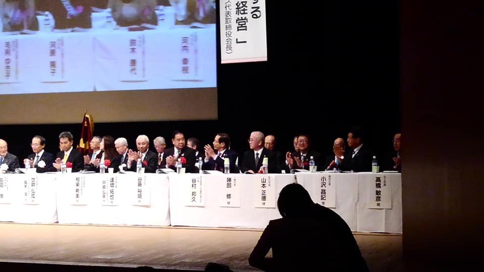 名前札が貼られたテーブルの席に着く、赤い胸章を着けた関係者の方々の写真