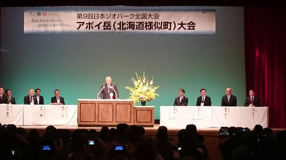 「第9回日本ジオパーク全国大会 アポイ岳(北海道様似町)大会」と書かれた横断幕が吊るされた舞台上で、左右に置かれたテーブルの席に着く関係者の方々と、中央に置かれた演台で話をする様子の男性の写真