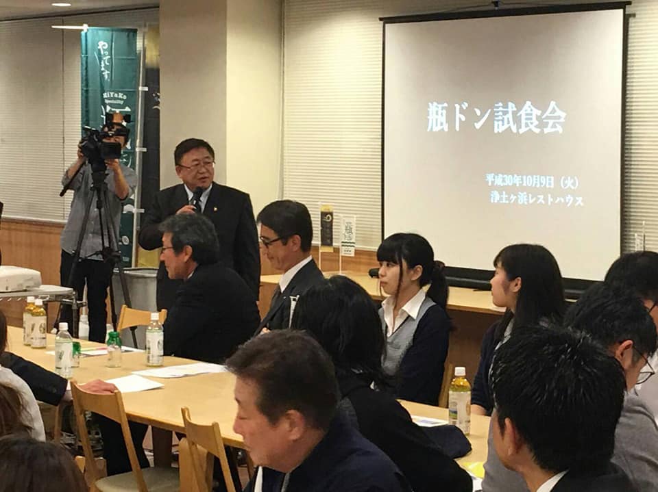 「瓶ドン試食会」という文字が写されたスクリーンが設置された会場で、テーブルに向か合うように座る参加者たちと、マイクを持って立ちながら話をする様子の市長の写真