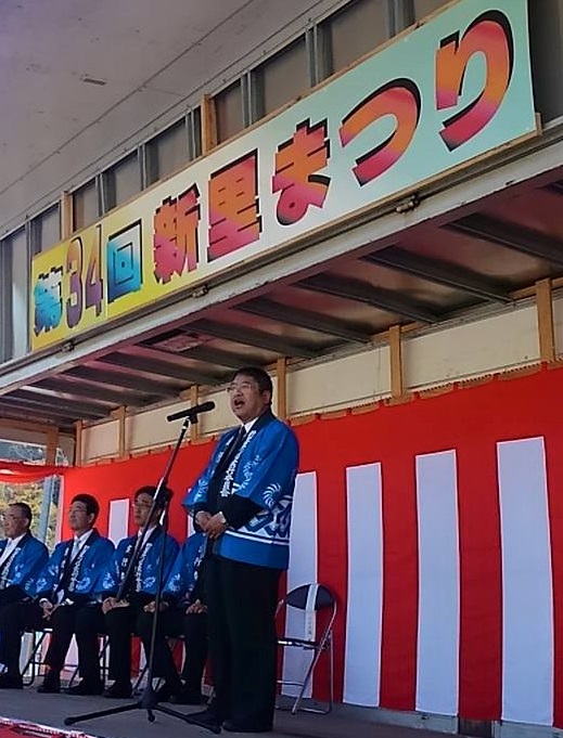 「第34回 新里まつり」と書かれた看板の下で、青い法被を着た市長がスタンドマイクに向かい話をする様子の写真