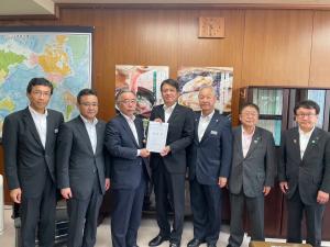 2枚の大きなポスターと世界地図が飾られた室内で、中央に農林水産省の角田秀穂政務官と市長会の代表が一緒に要望書を持って中央に立ち、市長を含む5名の関係者が横一列に並んでいる写真