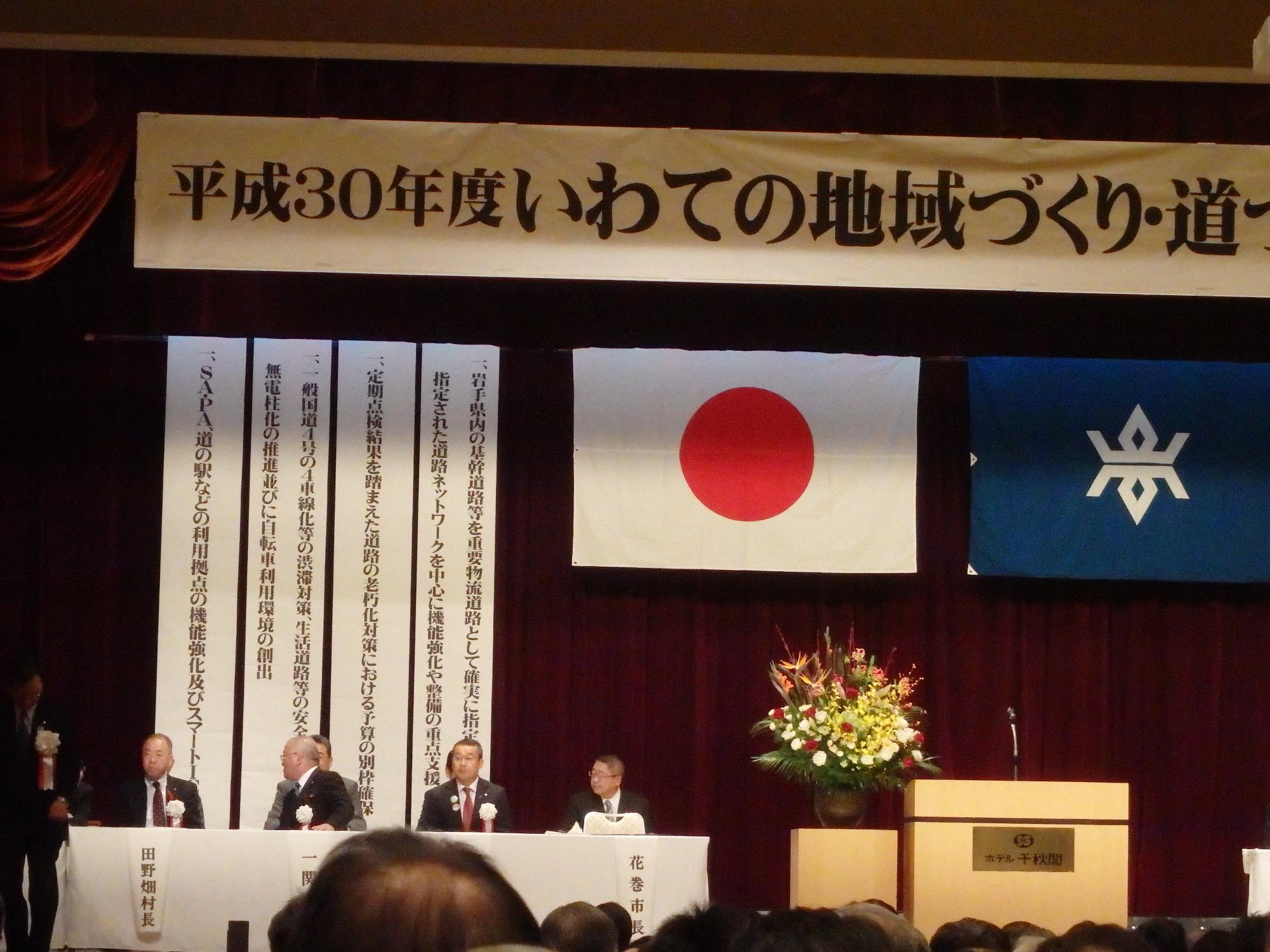 横断幕、日本国旗、垂れ幕などが吊るされた舞台上で、正面に名前札が貼られた白いテーブルクロスが敷かれた長机の席に、白い胸章をつけた市長と関係者の方々が座っている写真