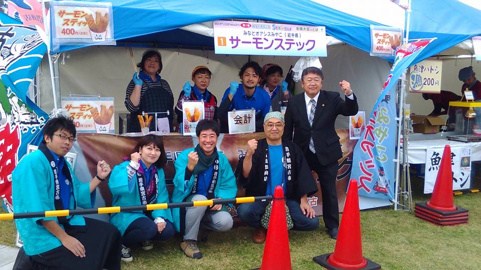 サーモンスティックを販売するテントで、市長と関係者の方々が握った左手の拳を顔の横に掲げてポーズをとっている記念写真