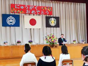「にゅうがくおめでとう」と書かれた幕、宮古市の市旗、国旗、校旗が貼られた体育館で、市長が座っている新1年生を前にして、演台で祝辞を述べている写真