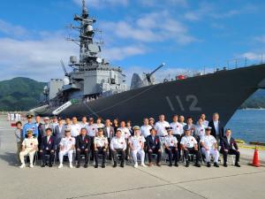停泊する海上自衛隊護衛艦「まきなみ」の前で、前列は椅子に座り、後列は立って横並びになっている関係者の方々の記念写真