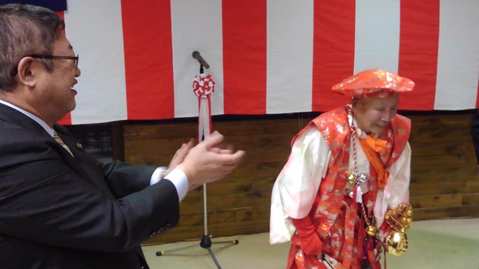 紅白幕が貼られた会場で、白い着物の上にオレンジ色の衣装を身にまとった荒田忠一さんがお辞儀をしている傍で市長が笑顔で拍手を送っている様子の写真