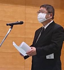 マイクスタンドの前で挨拶を行っている黒色のスーツを着用した市長をアップで撮影した写真