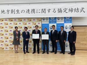 壁の上部に掲げられた「地方創生の連携に関する協定式」と書かれた横断幕と、壁際にバックパネルが置かれた会場で、締結書を正面に向けて両手で広げる男性と市長を中心に、左側に女性1名と男性1名、右側に男性3名が並んで写っている写真