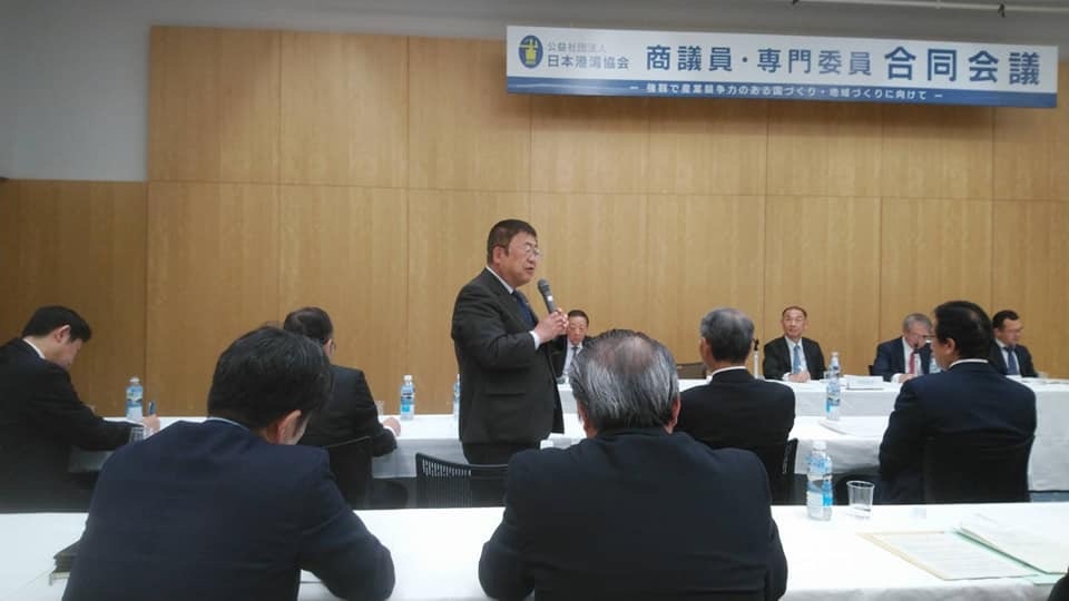 「日本港湾協会 商議員・専門委員会合同会議」と書かれた横断幕が掲げられた会場で、自席から立ち上がり右手にマイクを持って話をしている市長の写真