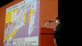 市長がステージのスクリーンに映し出された上空からの宮古港の交通ネットワークの向上について説明している写真
