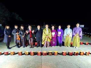 辺りが真っ暗な夜の遊覧船発着場で、ろうそくのがともされた灯篭のようなものが並べられ、その後方で色とりどりの袴や着物を着た方たちなど10名が横一列に並んでいる写真
